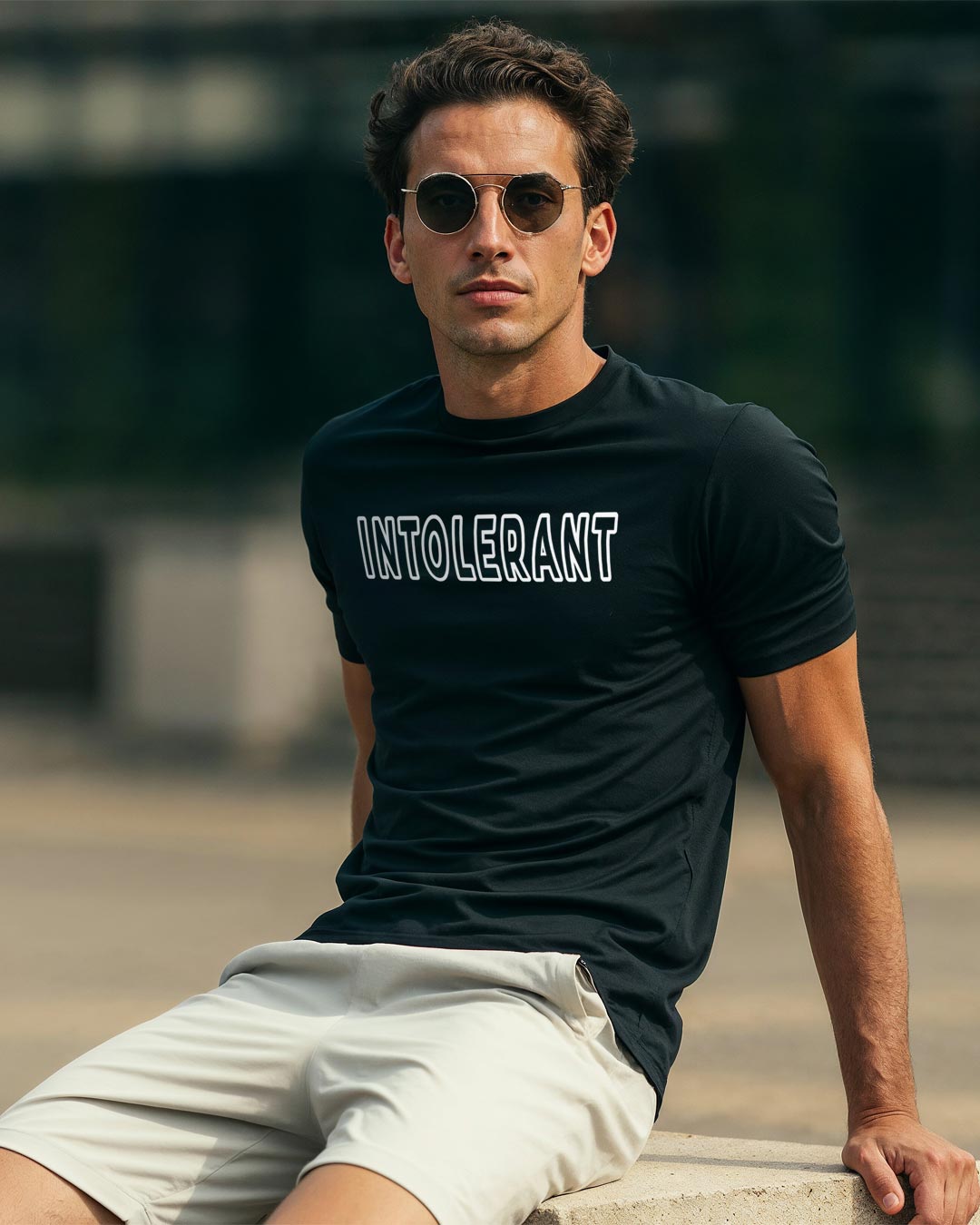 Ein junger Mann mit Sonnenbrille und schwarzem INTOLERANT Classic T-Shirt sitzt auf einem Stein in der Sonne.