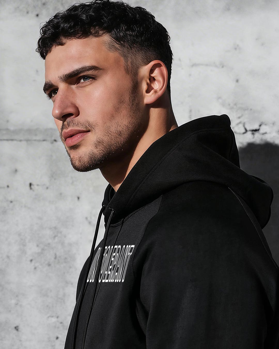 Ein junger Mann steht vor einer Betonwand und trägt einen schwarzen INTOLERANT Classic Hoodie.