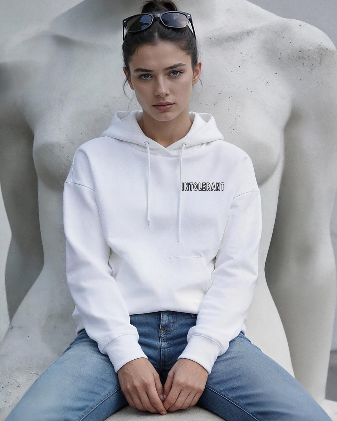 Weißer Kapuzenpullover der Brand INTOLERANT, getragen von einer Frau mit Sonnenbrille vor einer hellen Statue.