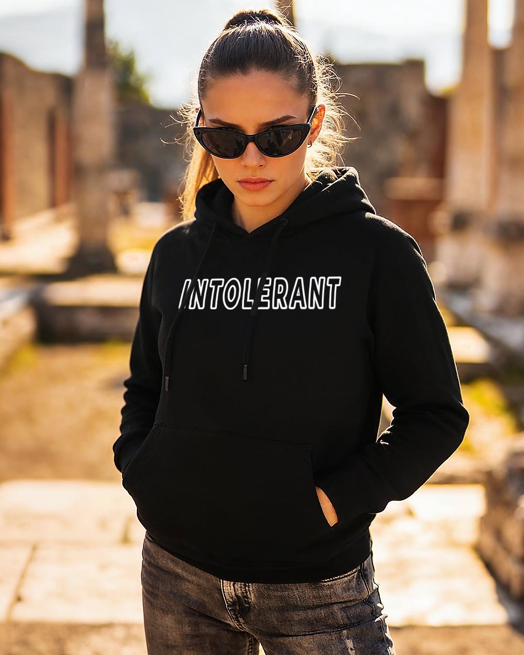 Junge Frau mit Sonnenbrille und Zopf vor antiken Steinruinen, trägt einen schwarzen INTOLERANT Classic Hoodie und eine Destroyed-Jeans in warmem Licht.