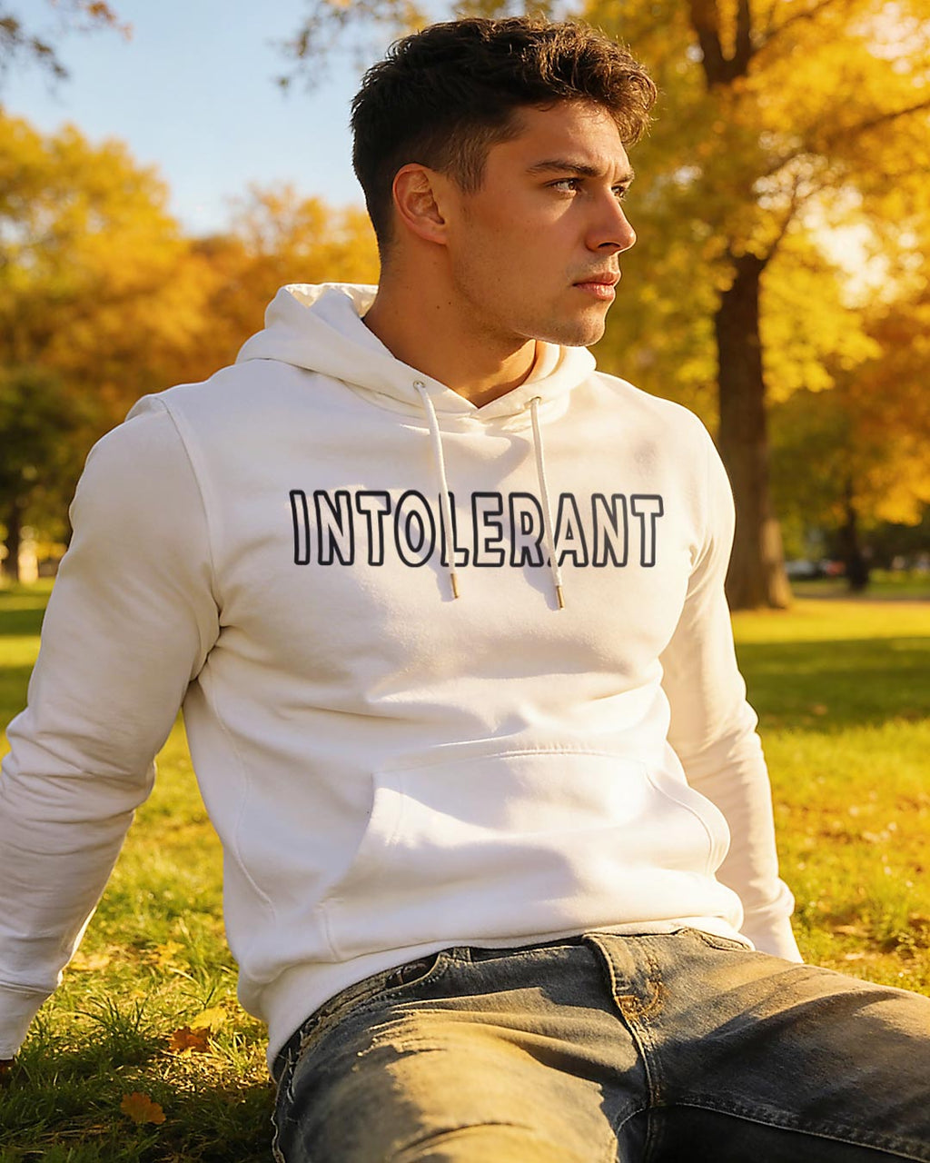 Hübscher junger Mann sitzt nachdenklich im Gras und stützt sich mit seinen Armen nach hinten ab. Er trägt einen weißen INTOLERANT Classic Hoodie und Bluejeans vor einer Parkkulisse in herbstlich warmem Sonnenlicht.