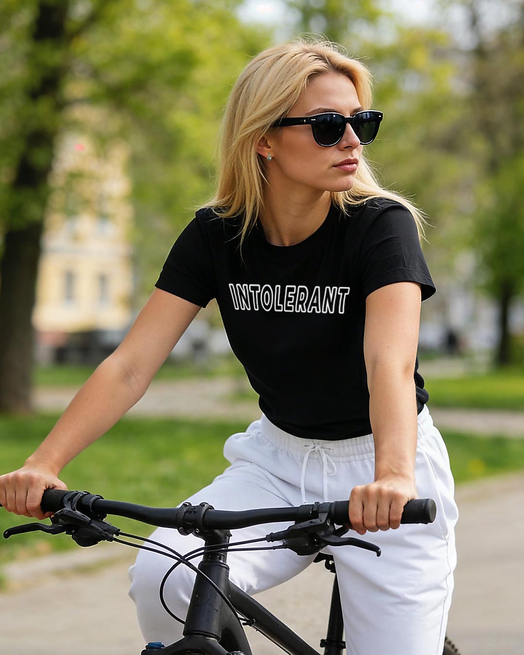 Eine junge blonde Frau auf einem Fahrrad, sie trägt eine Sonnenbrille, ein schwarzes INTOLERANT Classic T-Shirt und eine weiße Jogginghose. Im Hintergrund die Silhouette eines Stadtparks.