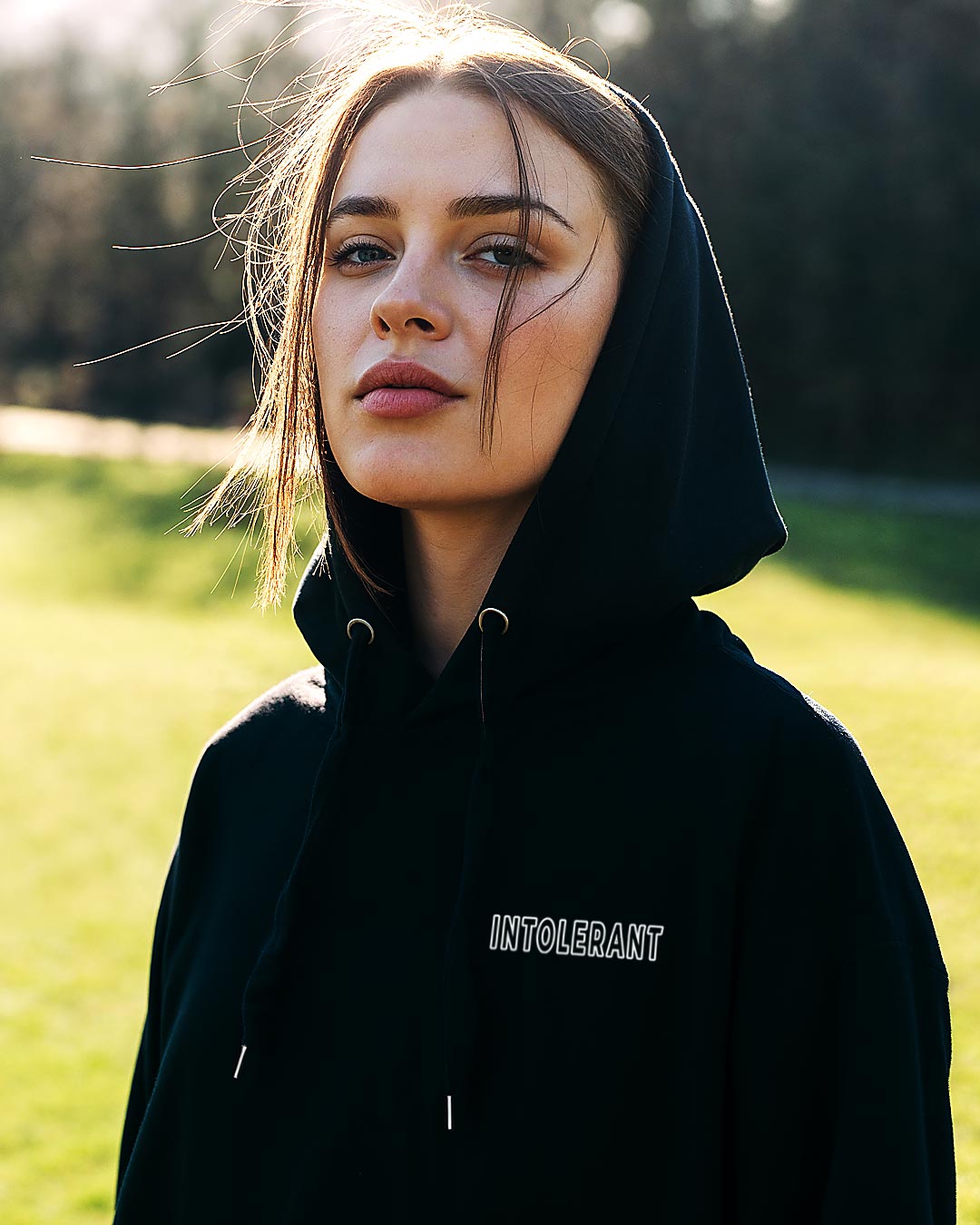 Nahaufnahme einer jungen Frau. Sie trägt einen schwarzen INTOLERANT Minimal Hoodie mit halb aufgesetzter Kapuze vor weichgezeichnetem, grünem Naturhintergrund bei sonnigem Licht.