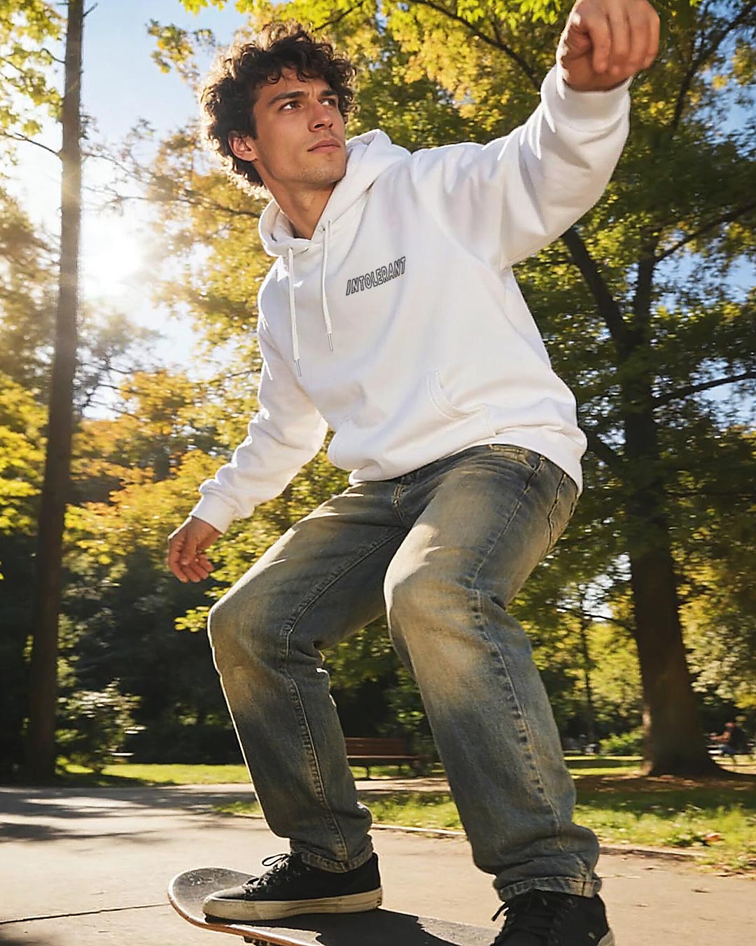 Junger Mann mit lockigem Haar beim Skateboarden im Park, trägt einen weißen INTOLERANT Minimal Hoodie und Bluejeans in sonniger, dynamischer Kulisse.