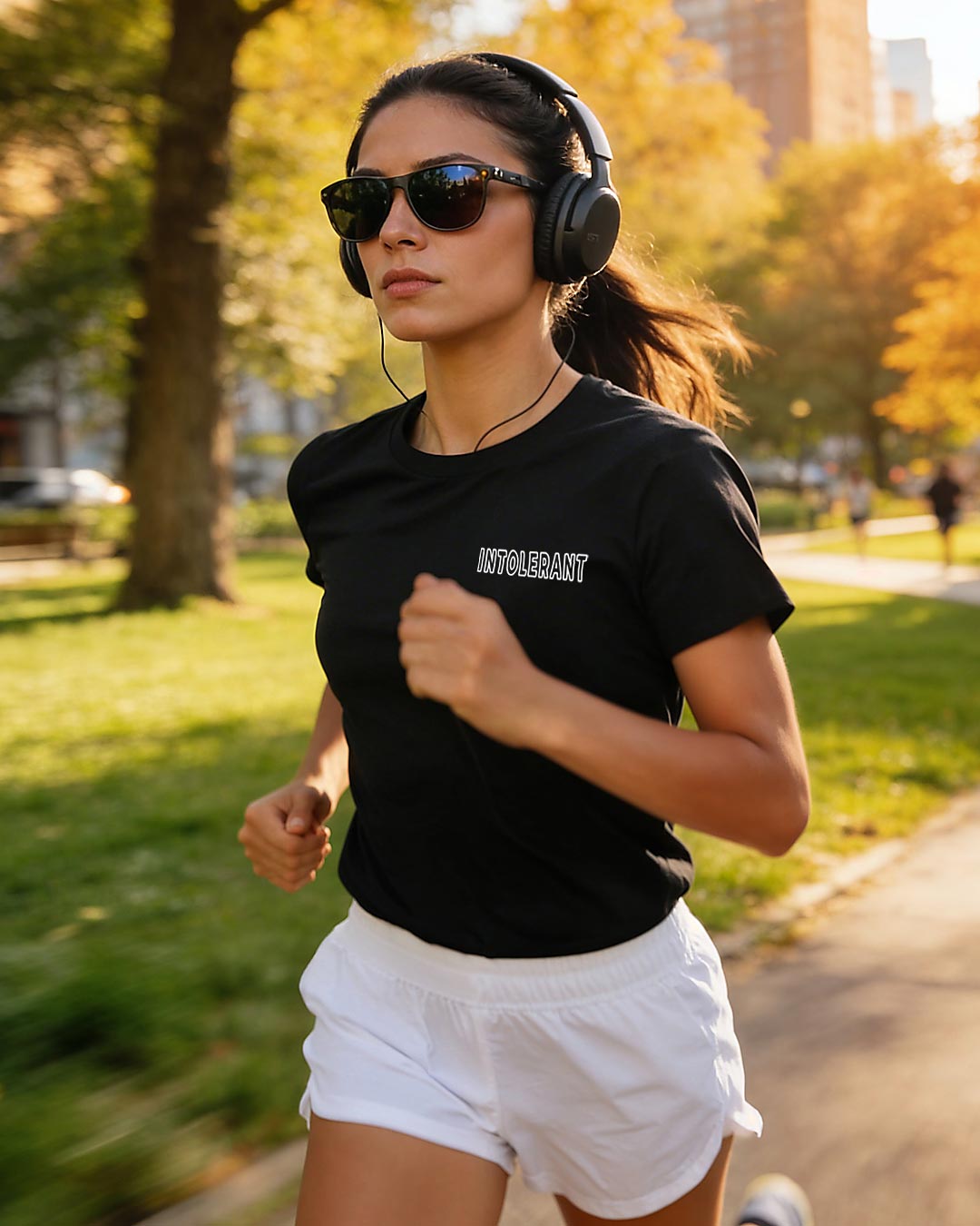 Frau beim Joggen im Park mit fokussiertem Blick, trägt ein schwarzes INTOLERANT Minimal T-Shirt, weiße Shorts, Sonnenbrille und Kopfhörer vor herbstlicher Kulisse im Gegenlicht.
