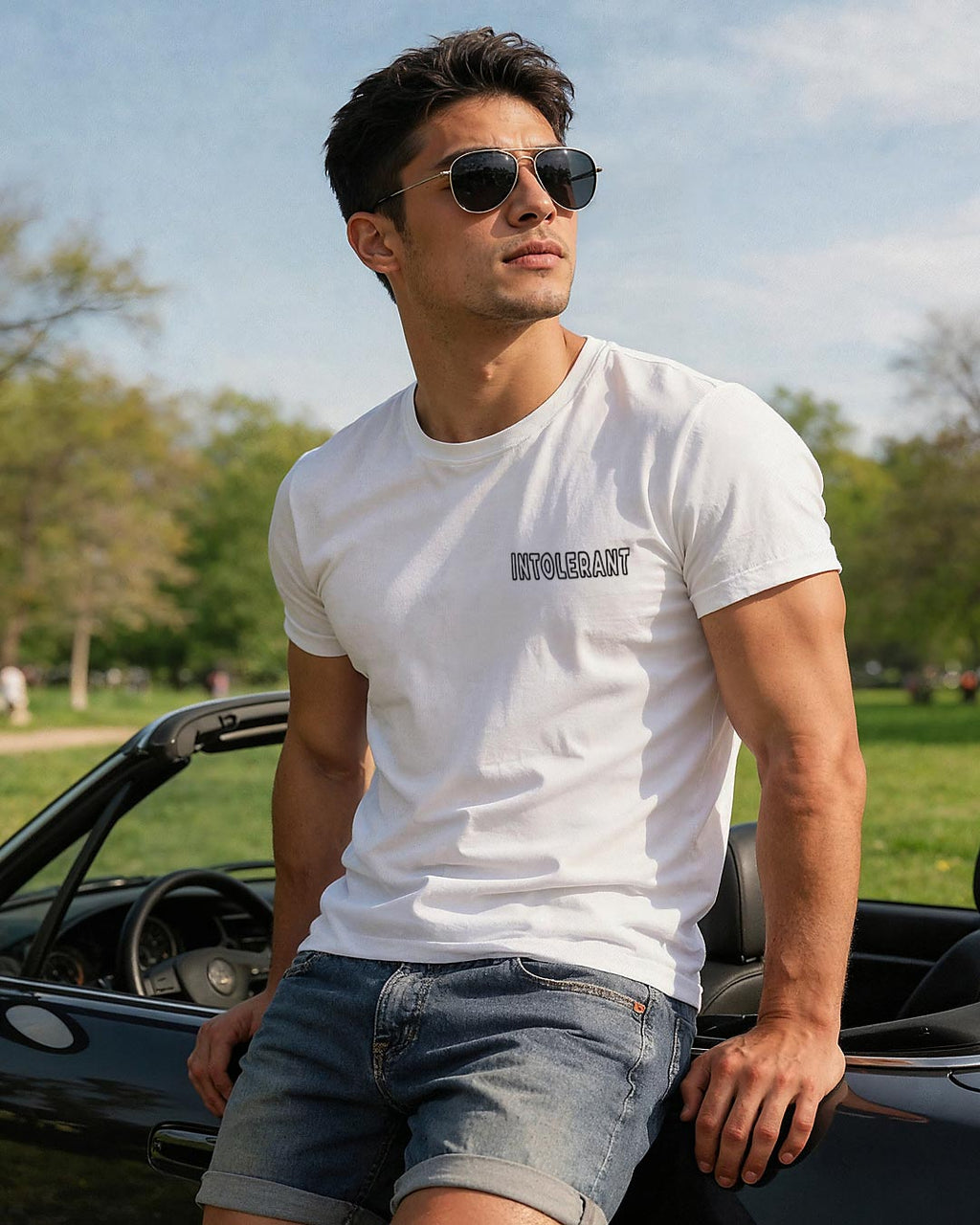 Junger Mann lehnt an einem Cabriolet vor einer Parkkulisse, trägt ein weißes INTOLERANT Minimal T-Shirt und eine kurze Jeans.